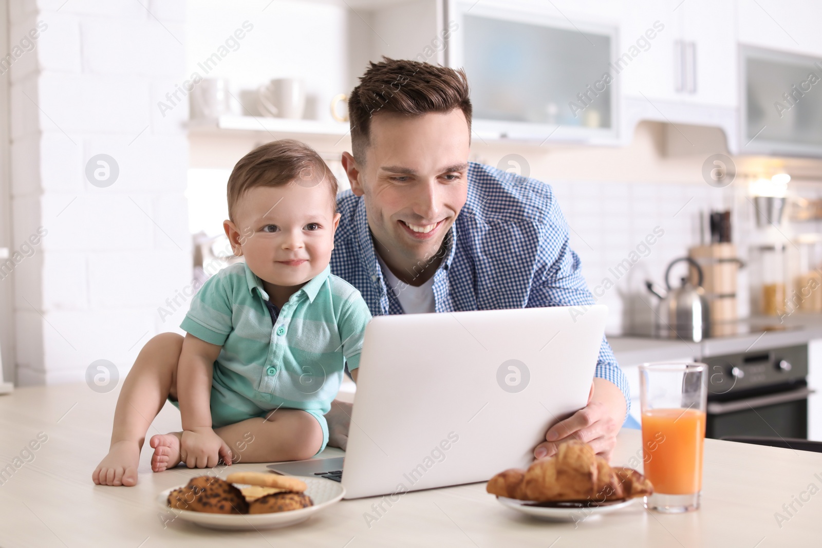 Photo of Young father with his cute little son using laptop in kitchen