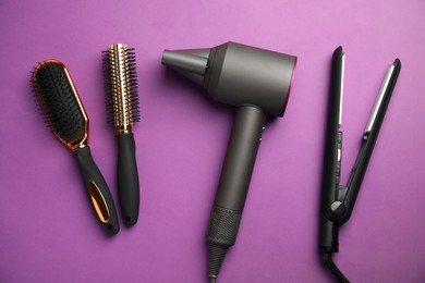 Hair dryer, straightener and brushes on purple background, flat lay