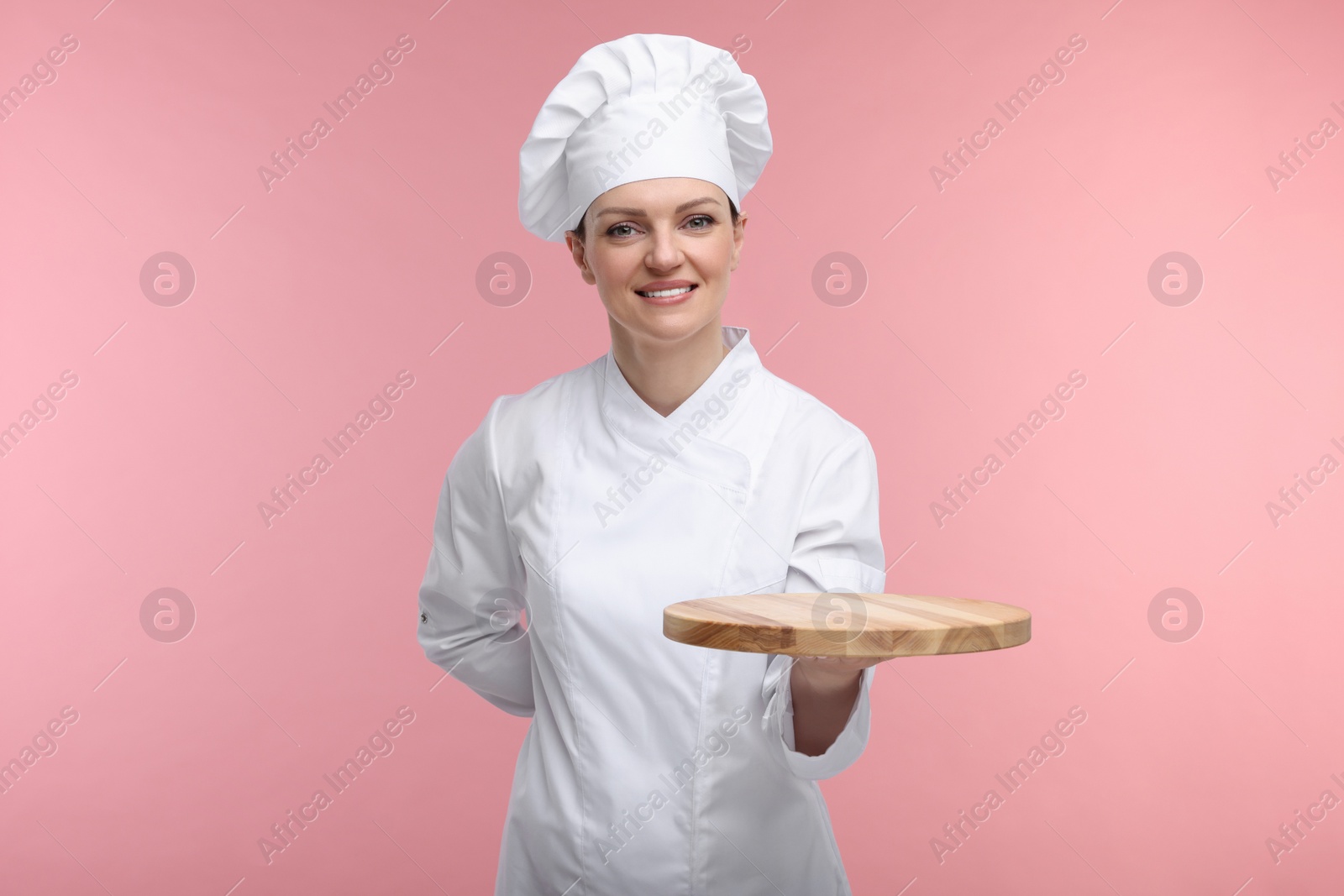 Photo of Happy chef in uniform holding empty wooden board on pink background