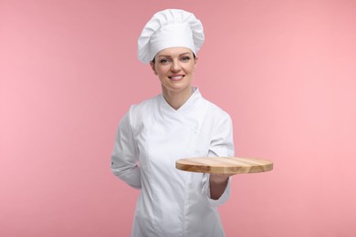 Photo of Happy chef in uniform holding empty wooden board on pink background