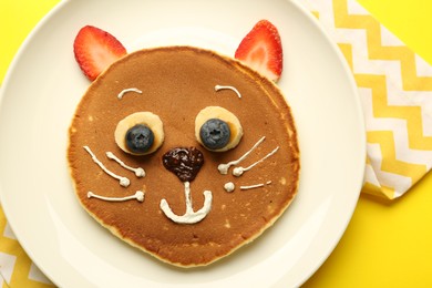 Photo of Creative serving for kids. Plate with cute cat made of pancakes, berries, cream, banana and chocolate paste on yellow background, top view