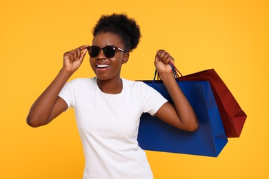 Happy young woman in stylish sunglasses with shopping bags on orange background