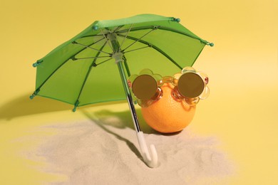 Funny orange with stylish sunglasses and umbrella on yellow background
