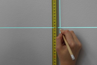 Woman using cross line laser level, tape and pencil for accurate measurement on grey wall, closeup