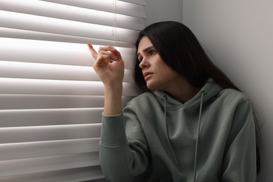 Photo of Sadness. Unhappy woman looking through window blinds at home