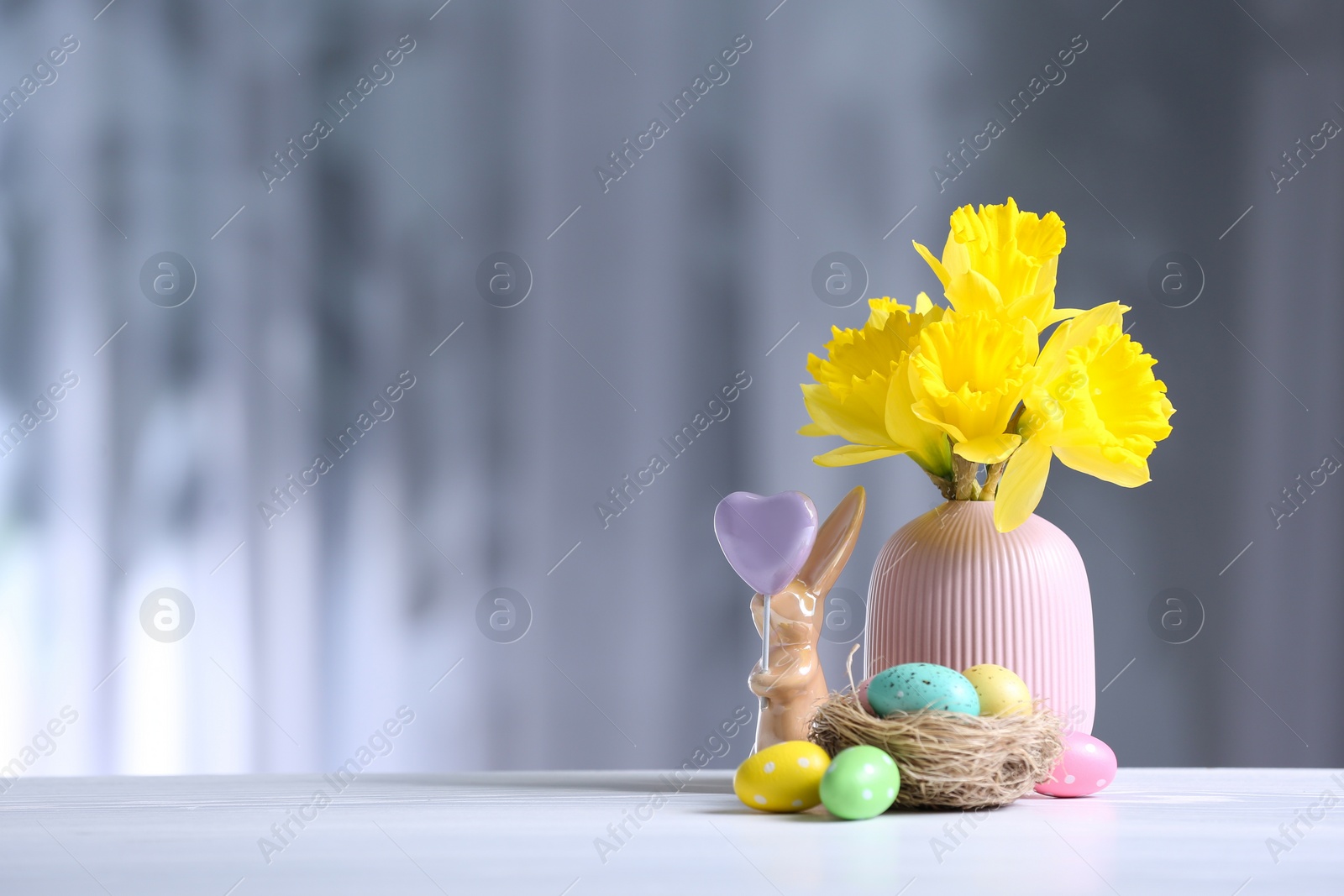 Photo of Painted Easter eggs, ceramic bunny and flowers on white table against blurred background. Space for text