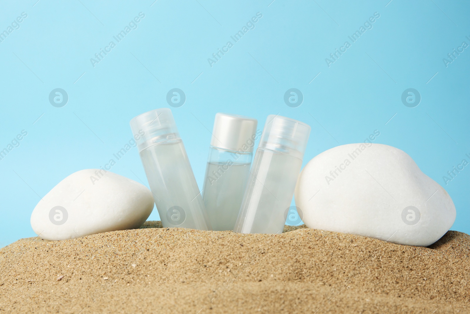 Photo of Bottles of serum and stones on sand against light blue background. Cosmetic product