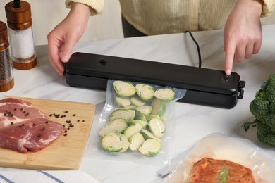Photo of Woman using sealer for vacuum packing with plastic bagzucchini at white table, closeup