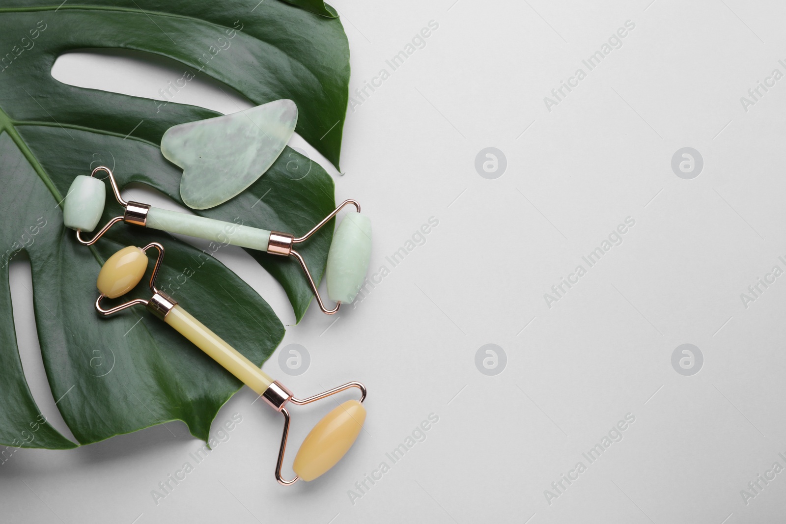 Photo of Gua sha stone, different face rollers and monstera leaf on light background, flat lay. Space for text