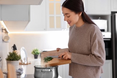 Photo of Smiling woman adding cut parsley into pot with soup in kitchen