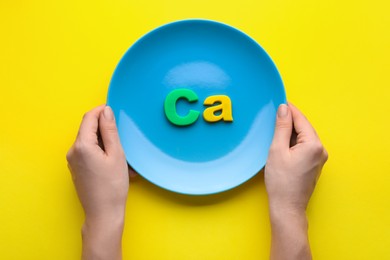 Woman holding plate with calcium symbol made of colorful letters on yellow background, top view