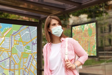 Image of Young woman in protective mask waiting for public transport at bus stop