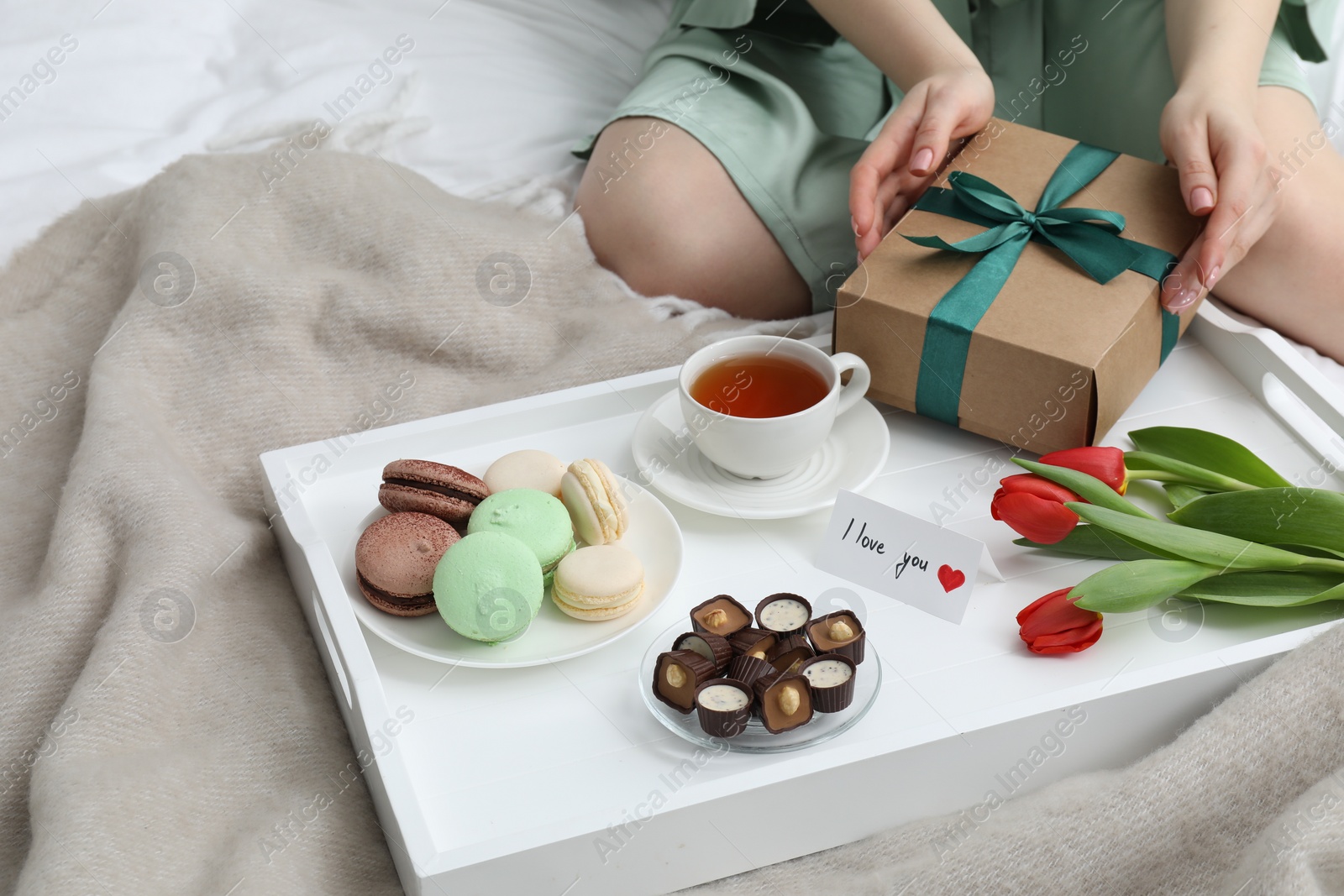 Photo of Tasty breakfast served in bed. Woman with gift box, desserts, tea, flowers and I Love You card at home, closeup