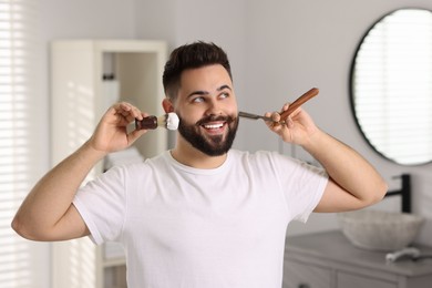 Handsome young man holding blade and shaving brush with foam in bathroom