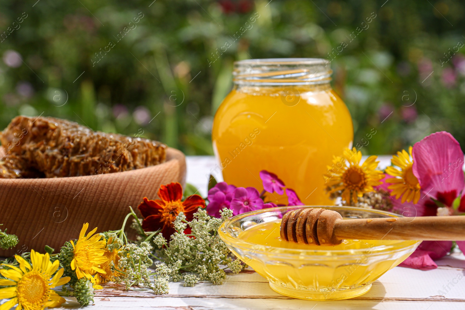 Photo of Delicious honey, combs and different flowers on white wooden table in garden