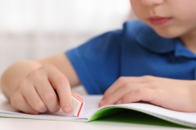 Photo of Little boy erasing mistake in his notebook at white desk indoors, closeup