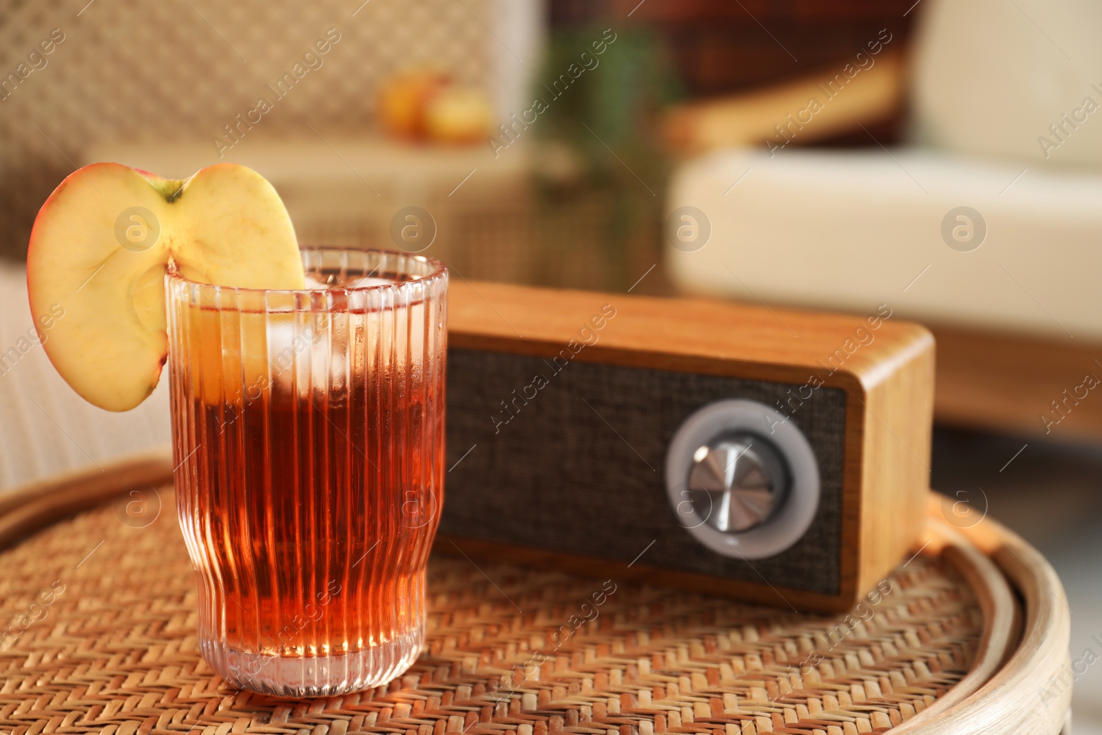 Photo of Relax at home. Glass of tasty cider and portable speaker on wicker table in room, closeup. Space for text