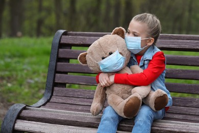 Little girl and teddy bear with protective masks on wooden bench outdoors. Space for text
