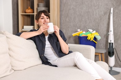 Beautiful young housewife with cup of drink resting after cleaning on sofa at home