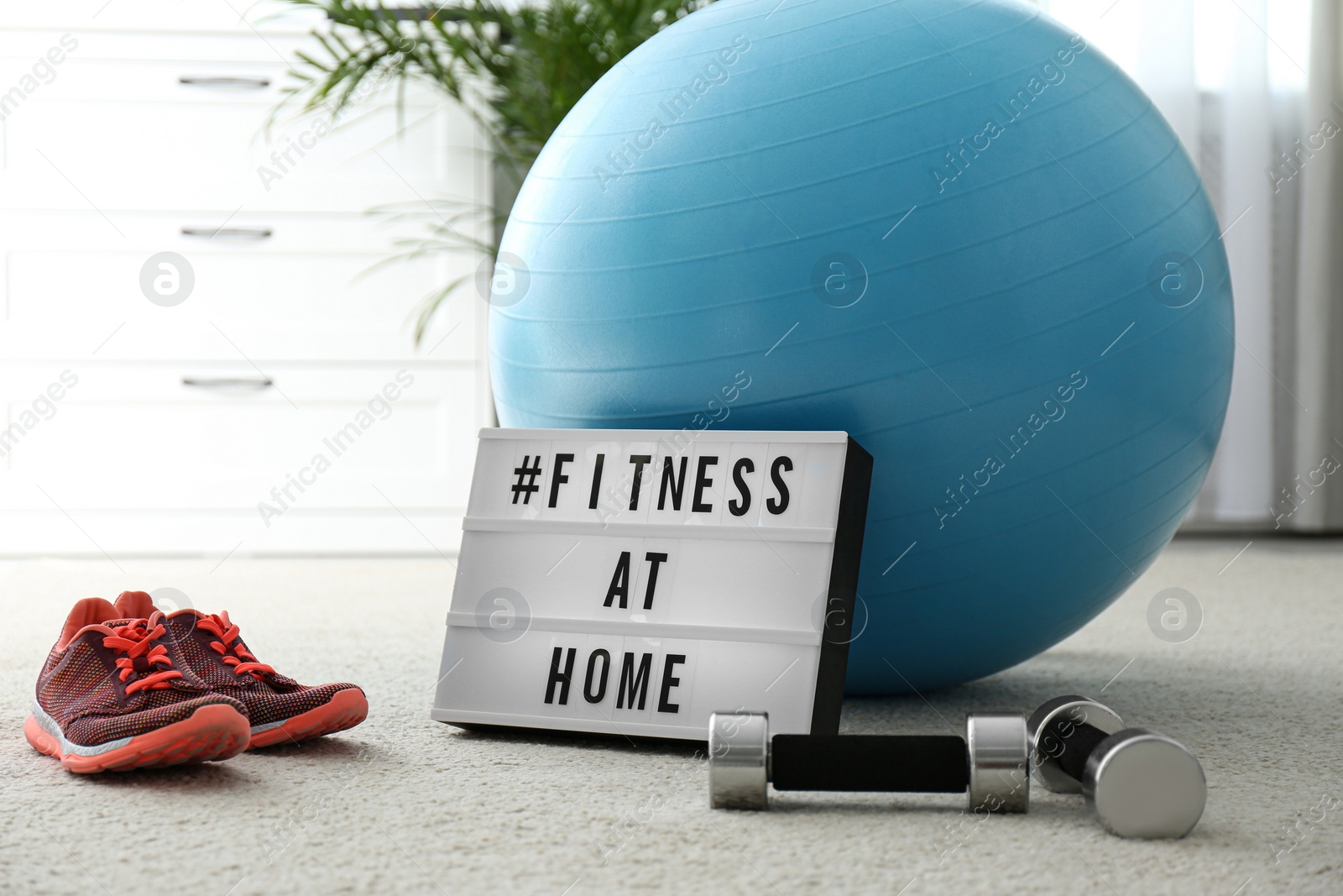 Photo of Sport equipment and lightbox with hashtag FITNESS AT HOME on floor indoors. Message to promote self-isolation during COVID‑19 pandemic