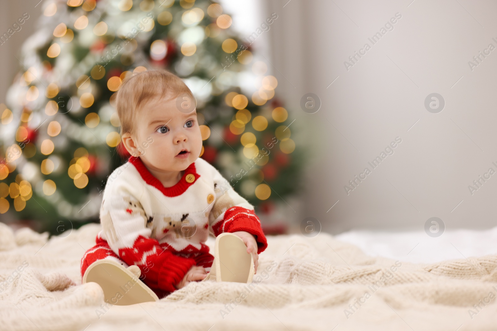 Photo of Cute little baby in Christmas sweater on knitted blanket against blurred festive lights, space for text. Winter holiday