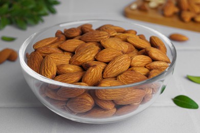 Bowl of delicious almonds and fresh leaves on white tiled table, closeup