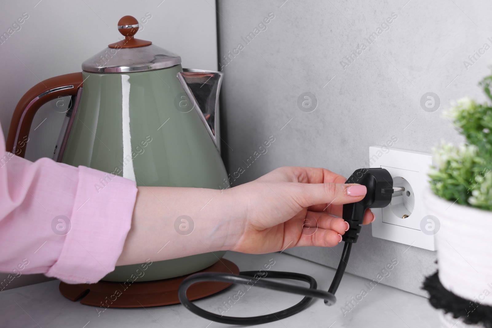 Photo of Woman plugging electric kettle into power socket at white table indoors, closeup