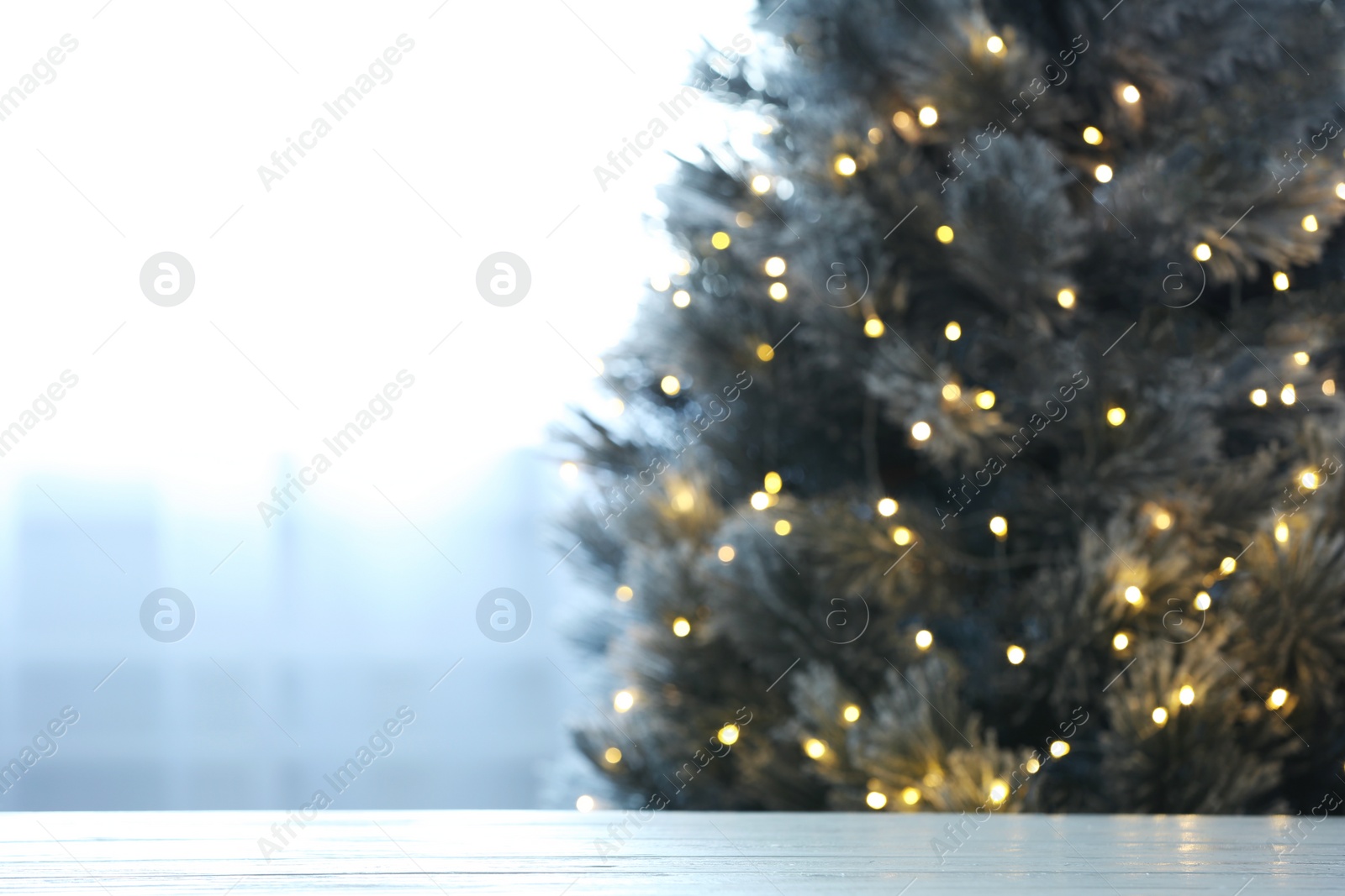 Photo of Blurred view of beautiful Christmas tree with yellow lights near window indoors, focus on wooden table. Space for text