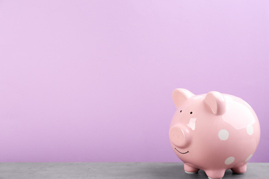 Pink piggy bank on light grey table against violet background. Space for text