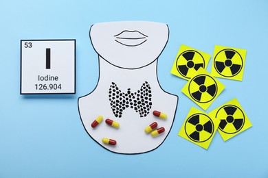 Photo of Card with chemical element Iodine, paper cutout of thyroid gland, radiation signs and pills on light blue background, flat lay