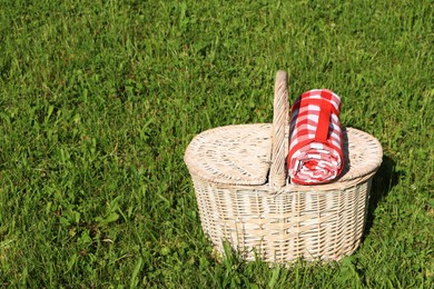 Rolled checkered tablecloth with picnic basket on green grass outdoors, space for text