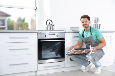 Photo of Young man holding baking sheet with cookies near oven in kitchen