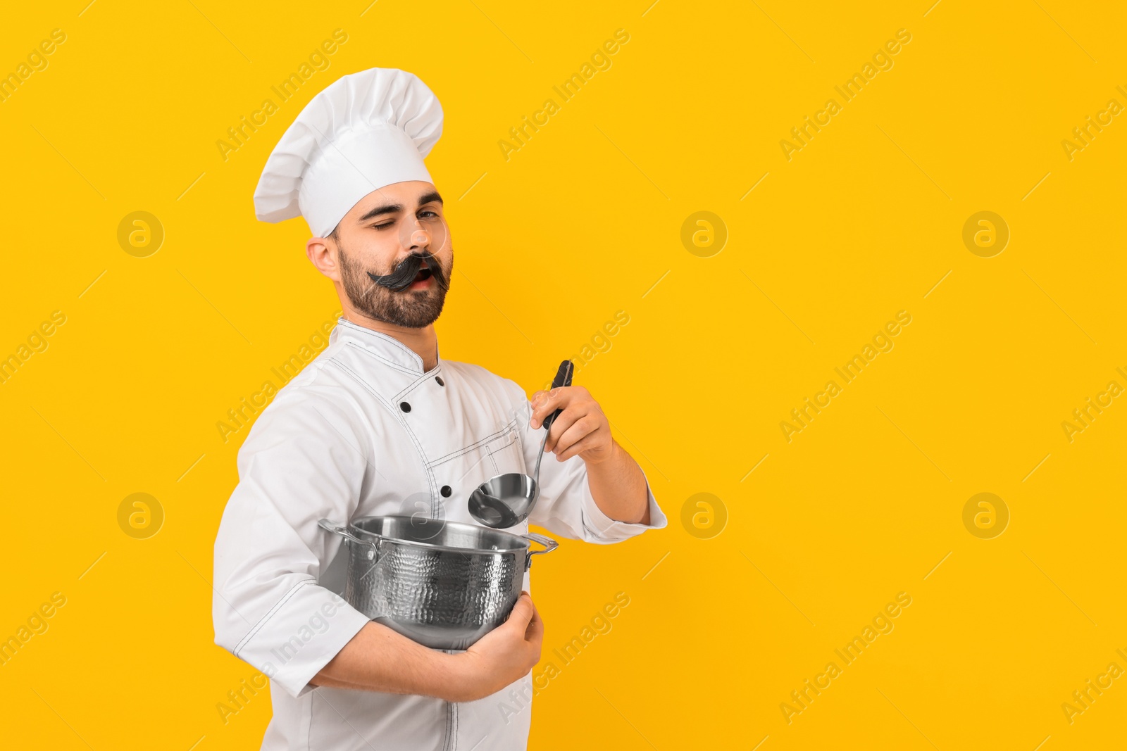 Photo of Professional chef with funny artificial moustache holding cooking pot and ladle on yellow background. Space for text