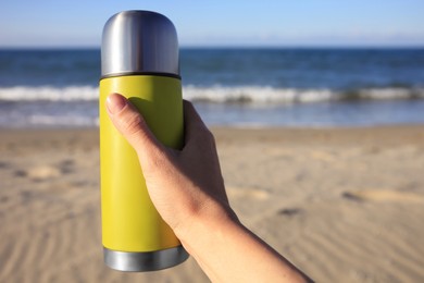 Photo of Woman holding yellow thermos with hot drink on beach near sea, closeup. Space for text