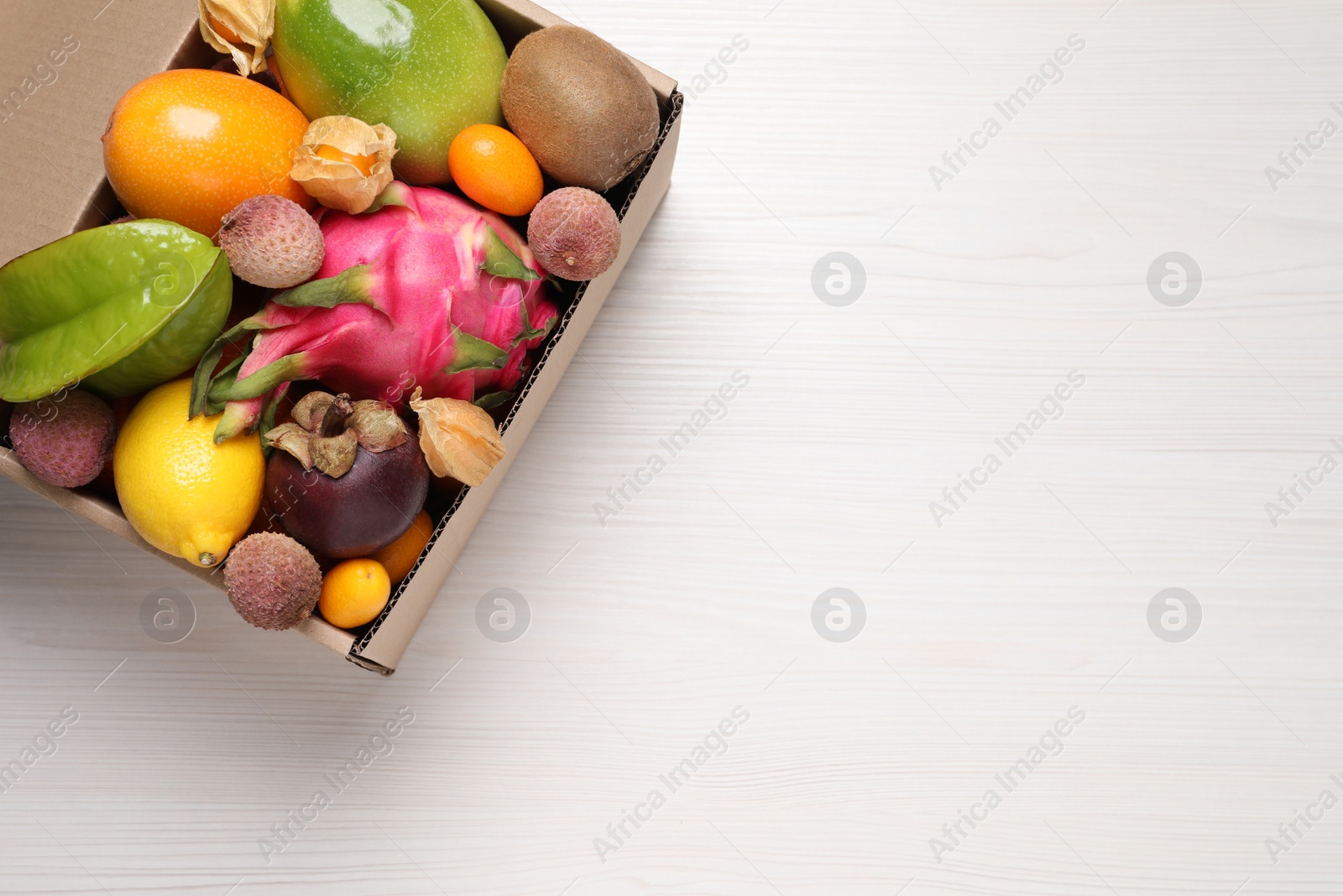 Photo of Cardboard box with different exotic fruits on white wooden table, top view. Space for text