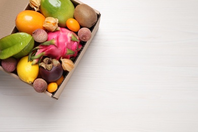 Photo of Cardboard box with different exotic fruits on white wooden table, top view. Space for text