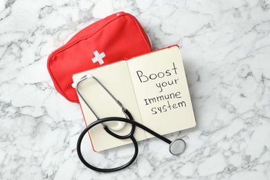 Photo of Notebook with words Boost Your Immune System, stethoscope and first aid kit on white marble table, flat lay