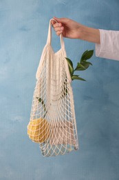 Photo of Woman holding net bag with different items against light blue wall, closeup. Conscious consumption