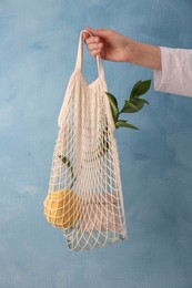 Woman holding net bag with different items against light blue wall, closeup. Conscious consumption