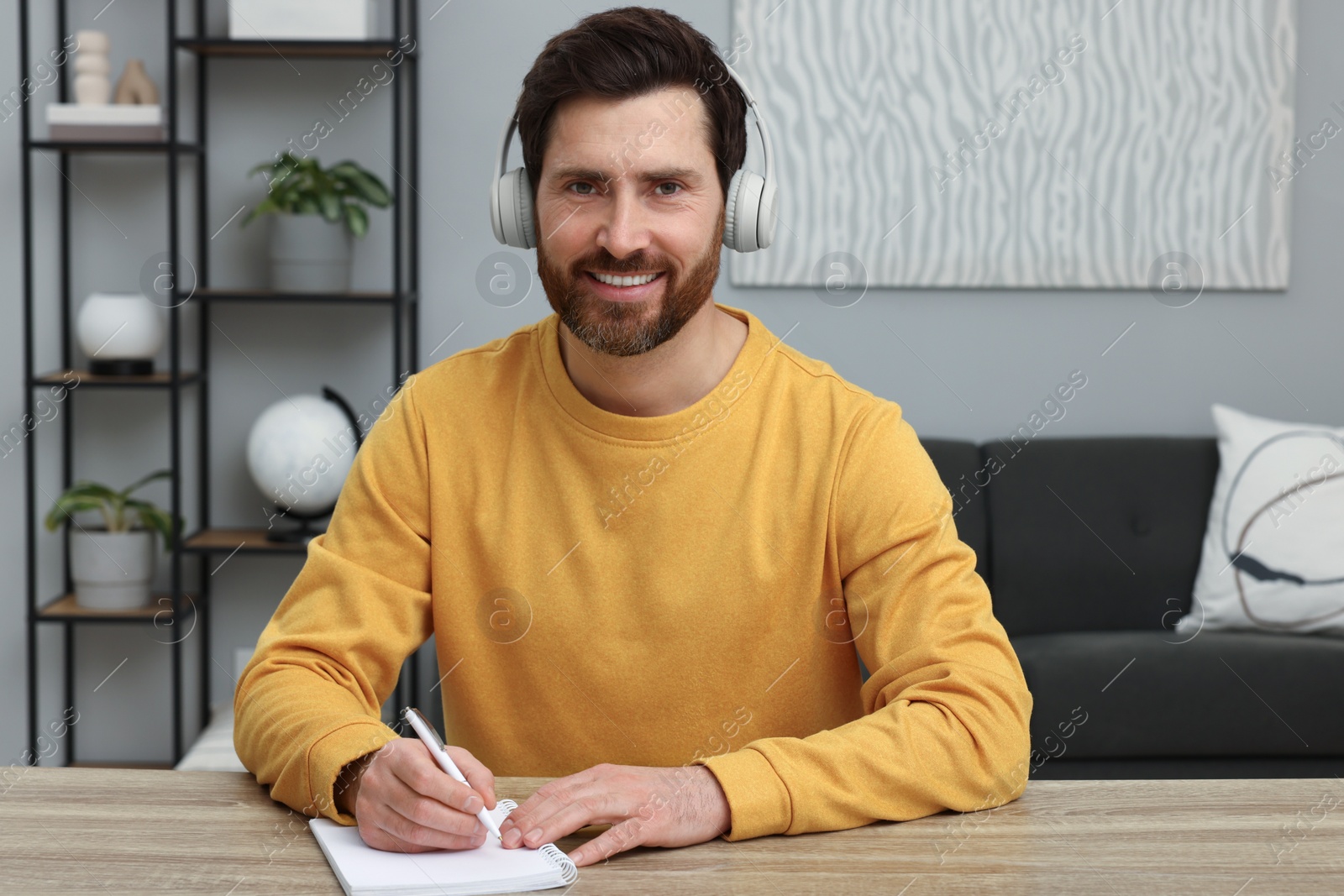 Photo of Man in headphones writing something into notebook at wooden table indoors