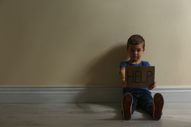 Little boy with sign HELP sitting on floor near yellow wall, space for text. Child in danger
