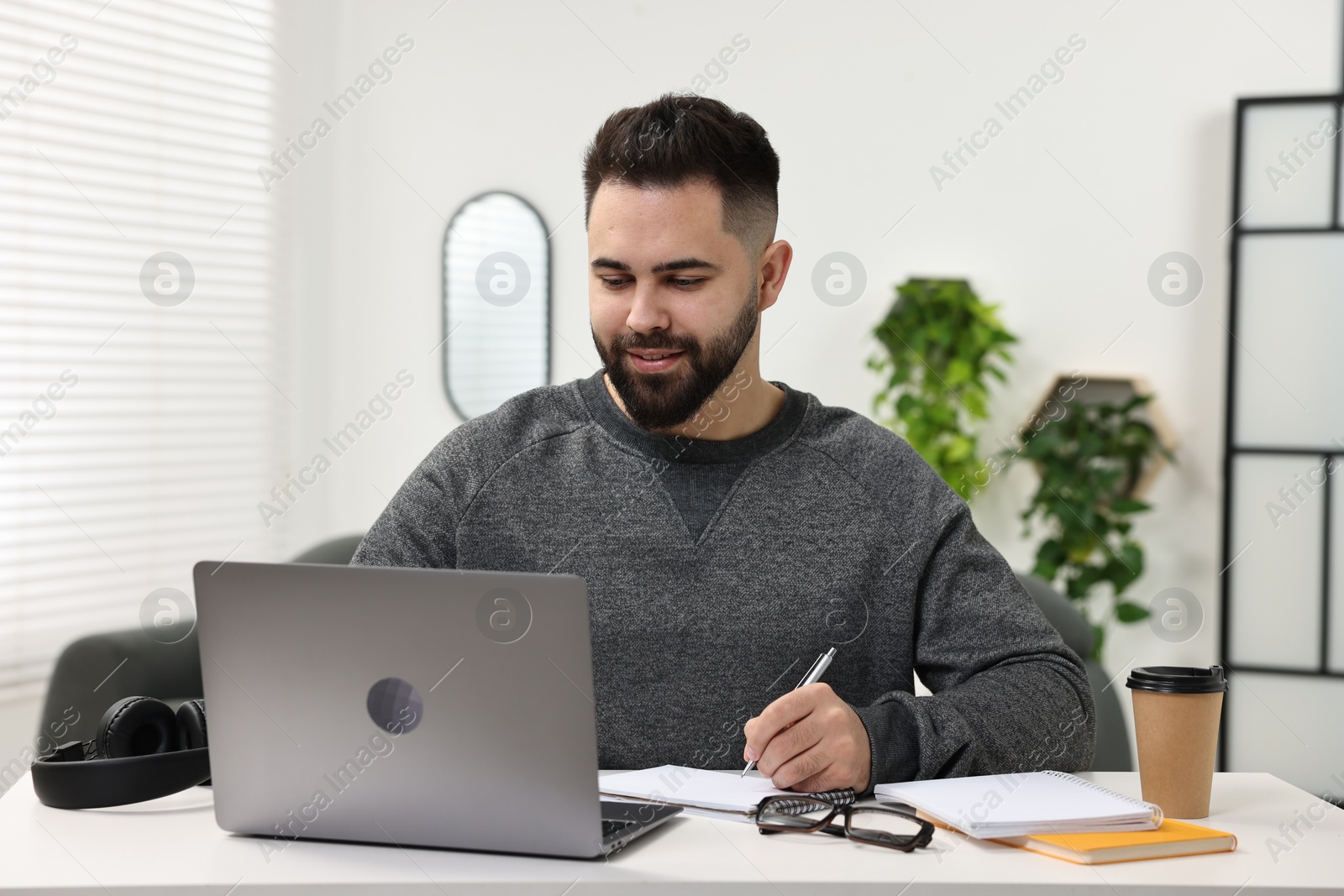Photo of E-learning. Young man taking notes during online lesson at white table indoors
