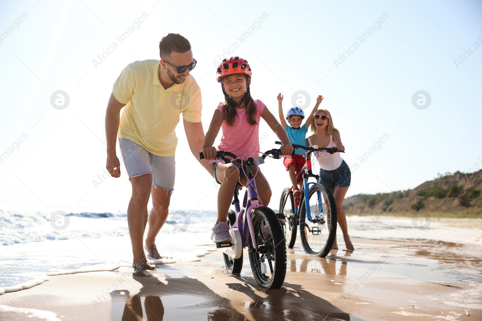 Photo of Happy parents teaching children to ride bicycles on sandy beach near sea