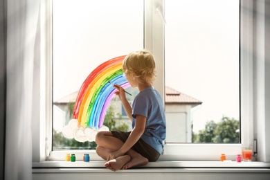 Photo of Little boy drawing rainbow on window indoors. Stay at home concept