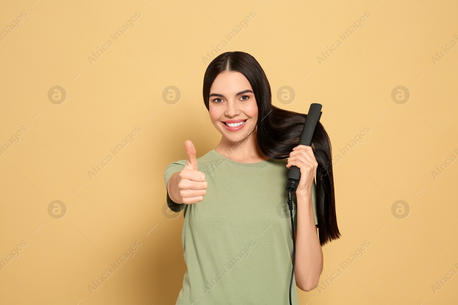 Photo of Beautiful happy woman showing thumbs up while using hair iron on beige background