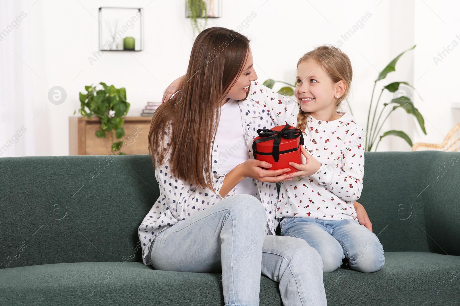 Photo of Cute little girl presenting her mother with gift on sofa at home