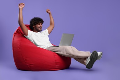 Photo of Happy man with laptop sitting on beanbag chair against purple background