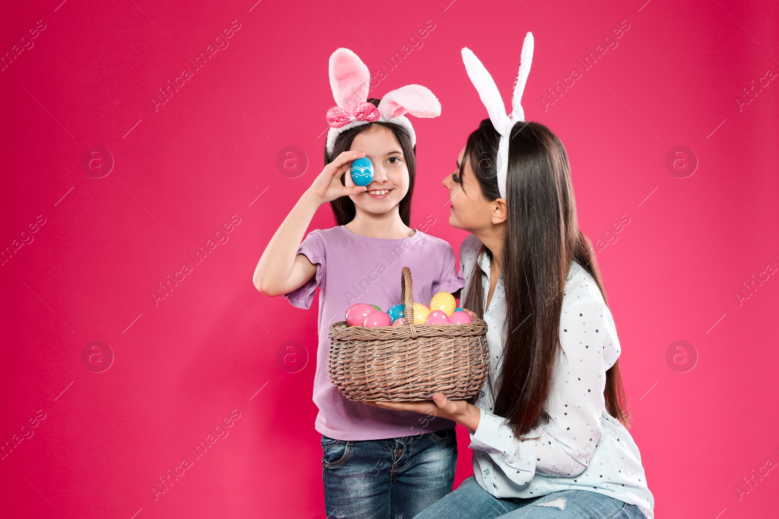 Photo of Mother and daughter in bunny ears headbands with basket of Easter eggs on color background