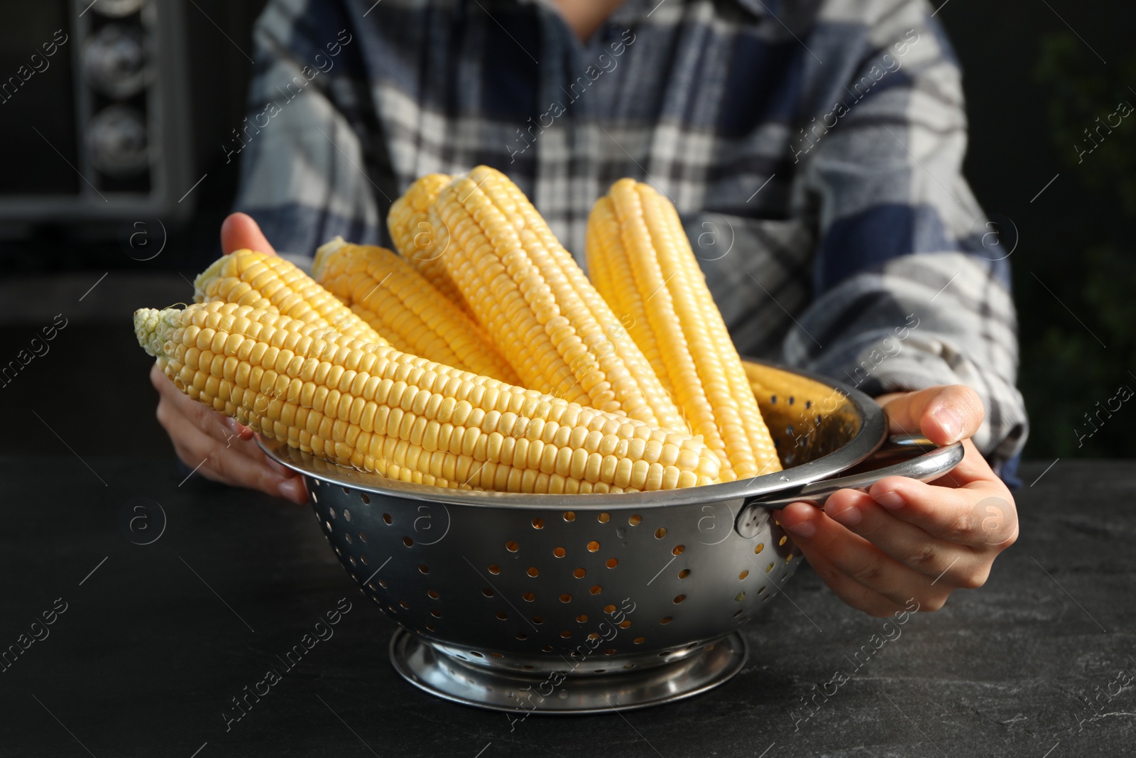 Photo of Woman with colander of corn cobs at black table, closeup