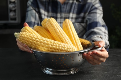 Photo of Woman with colander of corn cobs at black table, closeup